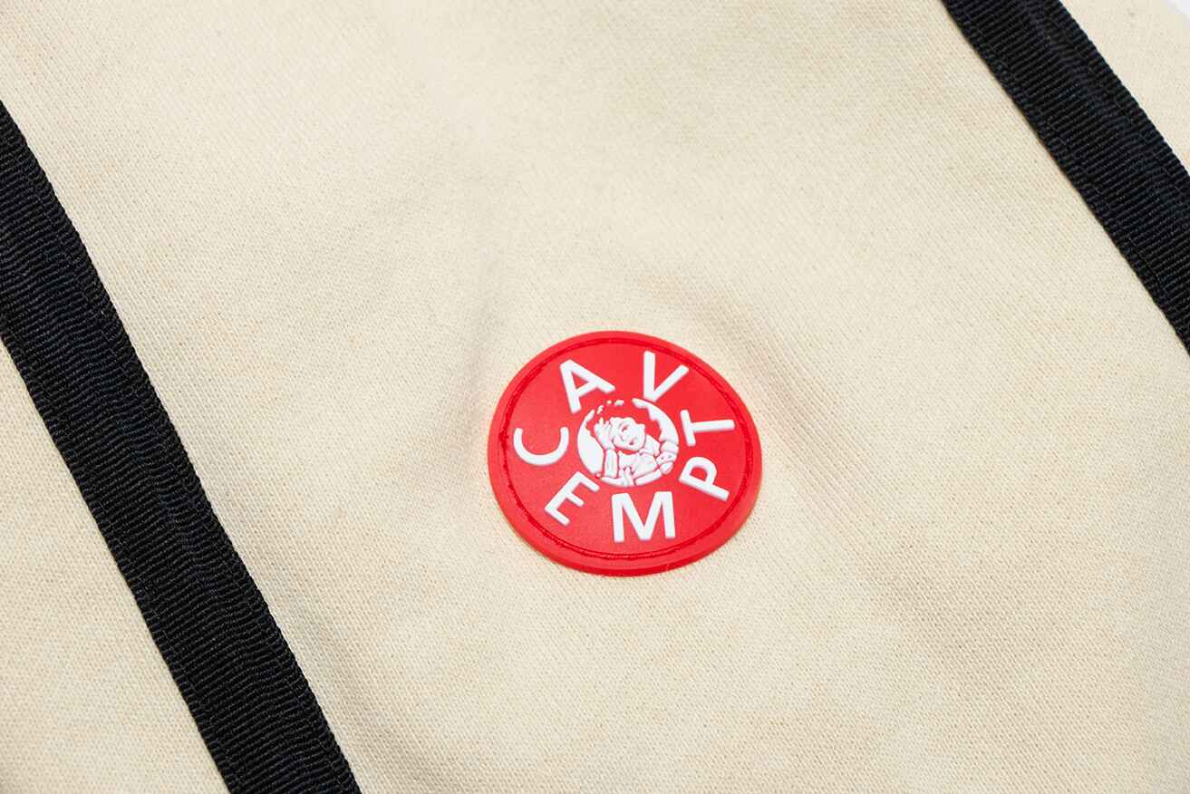 Soft beige backpack with black straps and red circular badge featuring a cartoon character and the words "CAV EMPT" on it.